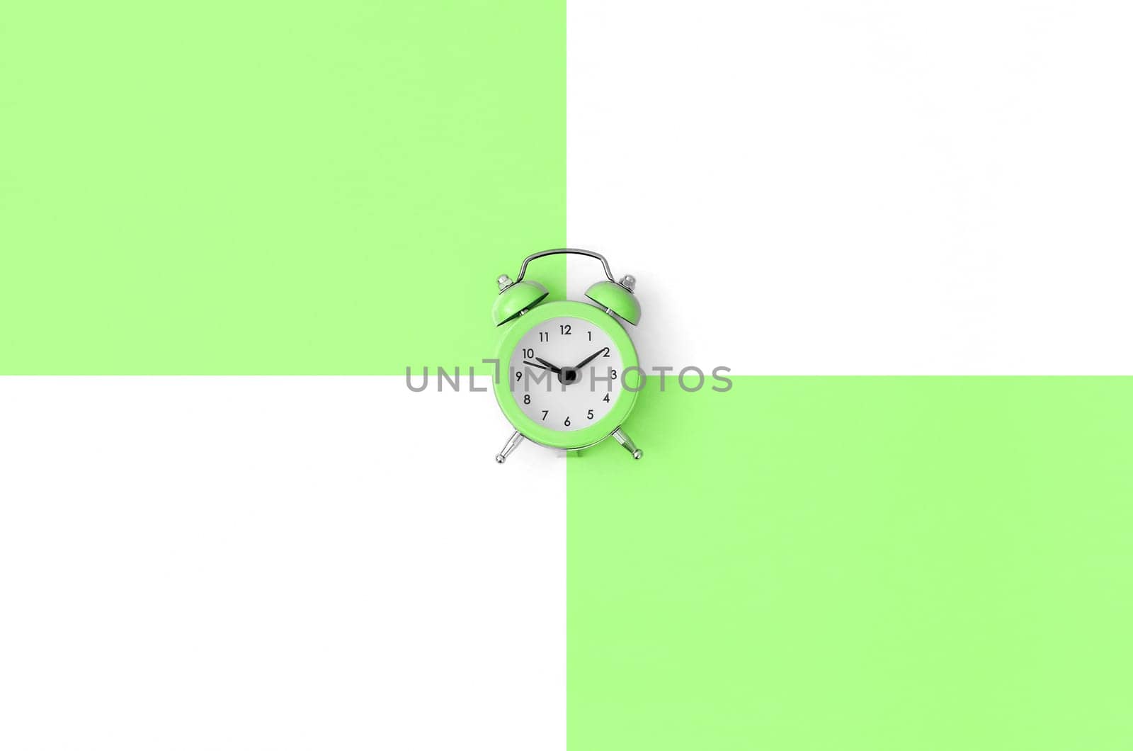 A small green alarm clock is located between areas of green and white colors. Time management concept. Flat lay minimal template with copy space. Top view