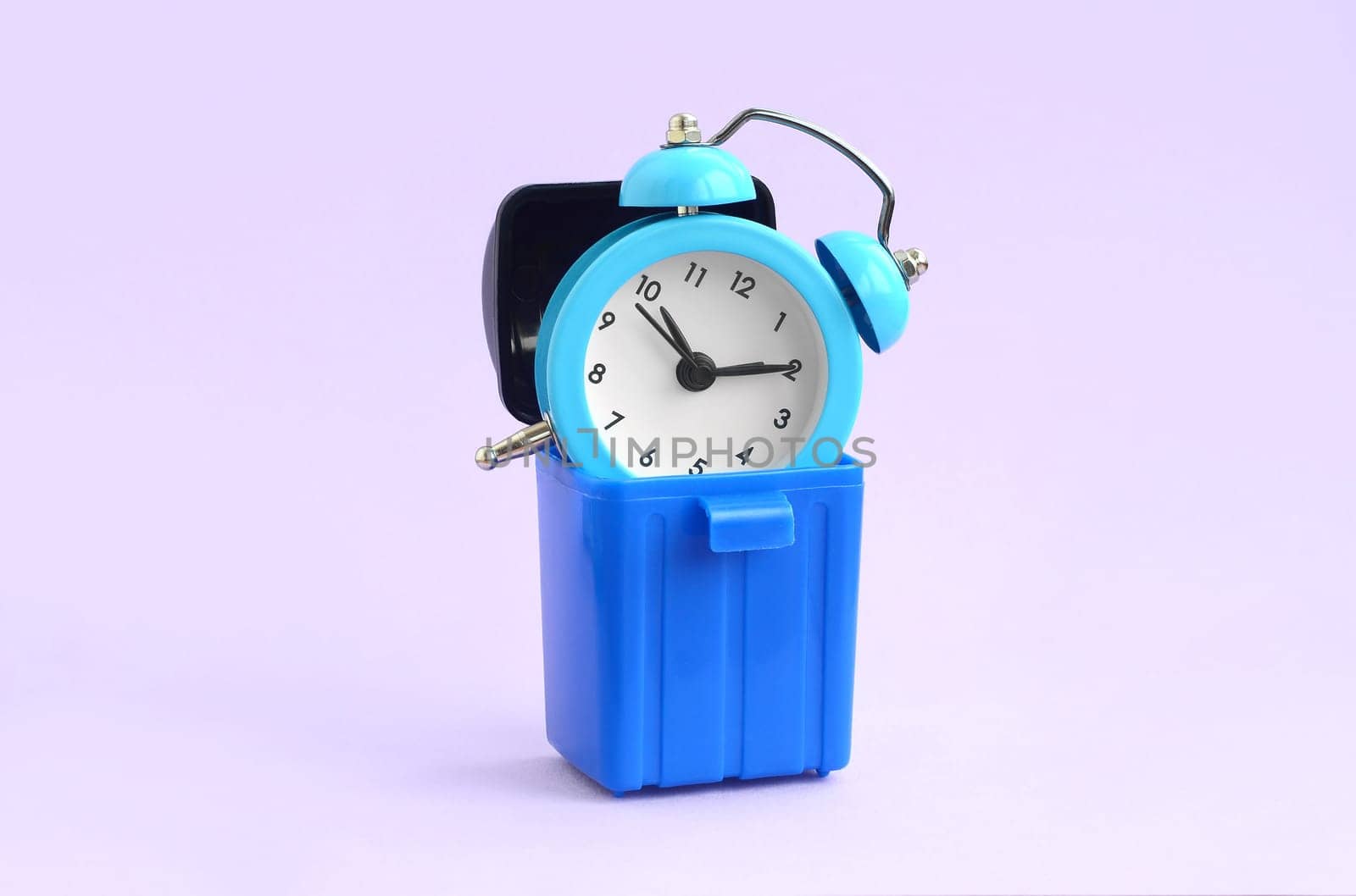 Wasting time concept. Blue alarm clocks in plastic trash bin on violet background. Bad and wrong time management