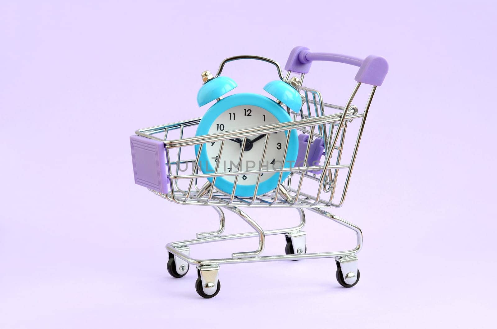 Blue alarm clock in supermarket trolley on violet background. Time resource acquisition concept. Time to sale
