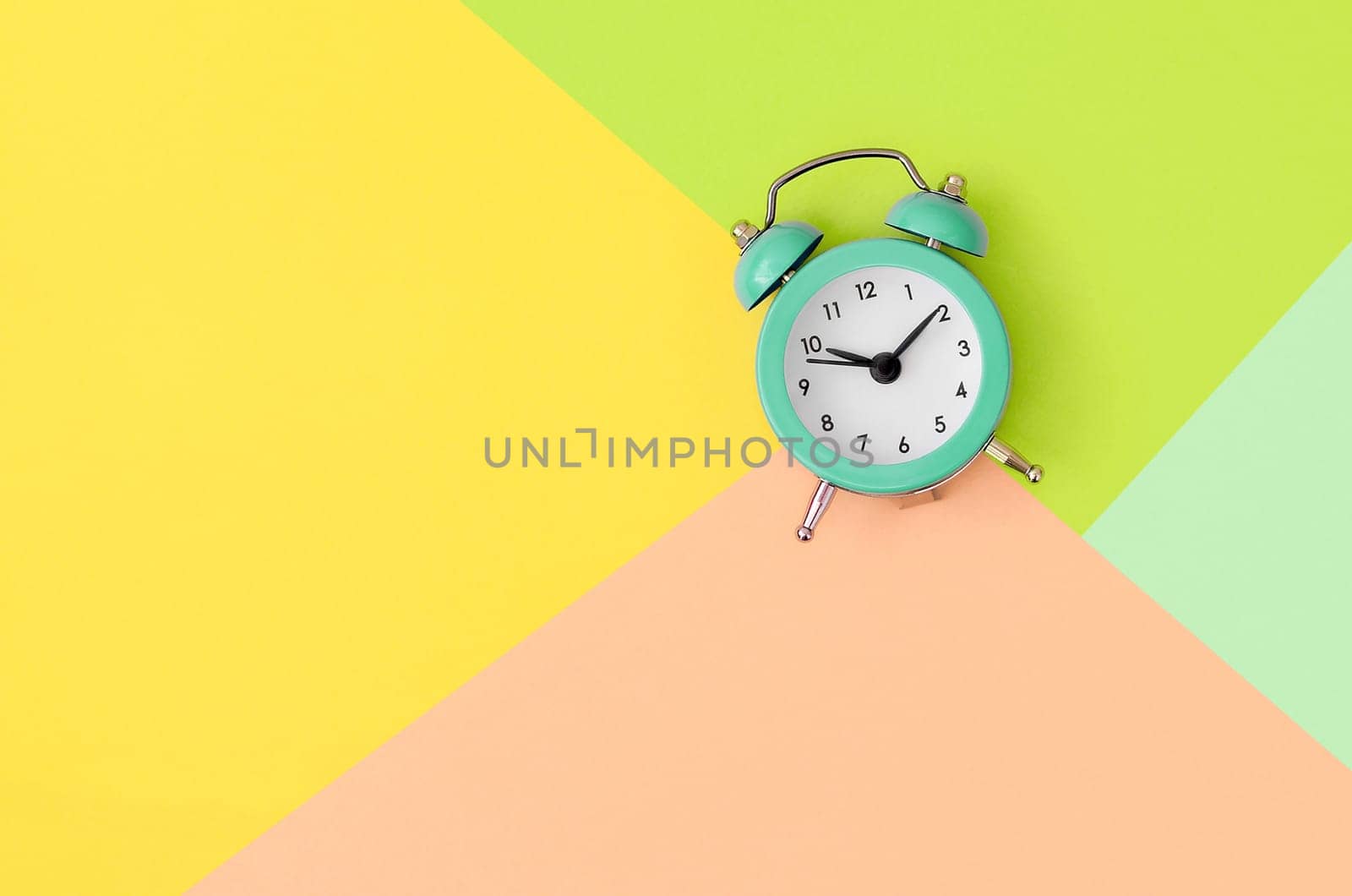 A small turquoise alarm clock is located between areas of yellow, peach and green colors. Time management concept. Flat lay minimal template with copy space. Top view