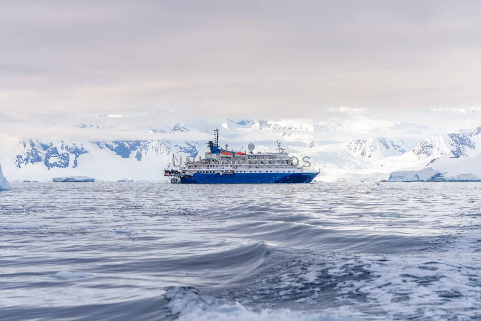 a blue, small expedition ship lies in front of an Antarctic iceberg landscape in Cierva Cove - a deep inlet on the west side of the Antarctic Peninsula, surrounded by rugged mountains and dramatic glacier fronts.