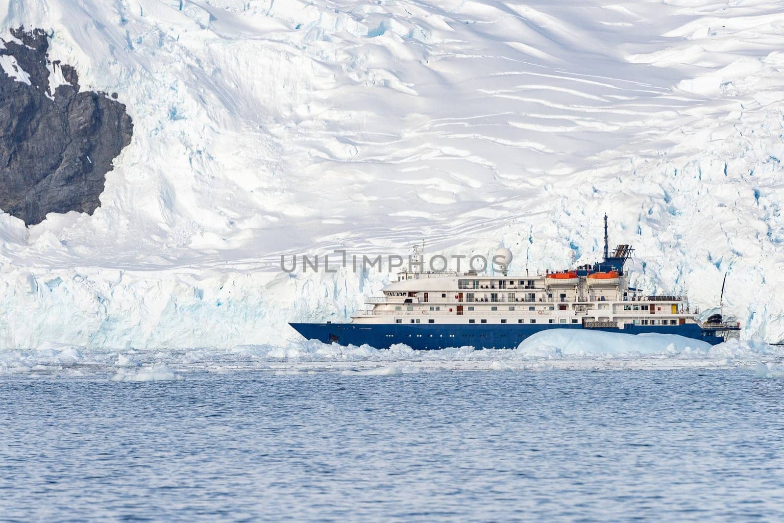a blue, small expedition ship lies in front of an Antarctic iceberg landscape in Cierva Cove - a deep inlet on the west side of the Antarctic Peninsula, surrounded by rugged mountains and dramatic glacier fronts.
