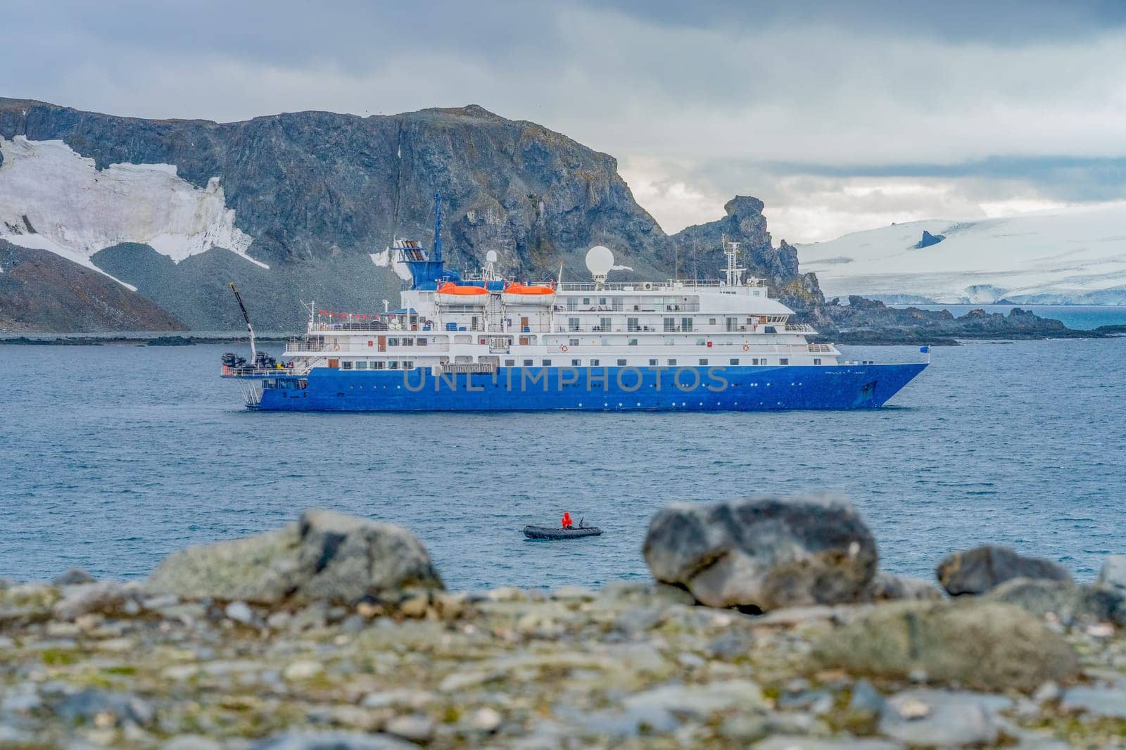a blue, small expedition ship lies in front of an Antarctic iceberg landscape in Cierva Cove - a deep inlet on the west side of the Antarctic Peninsula, surrounded by rugged mountains and dramatic glacier fronts.