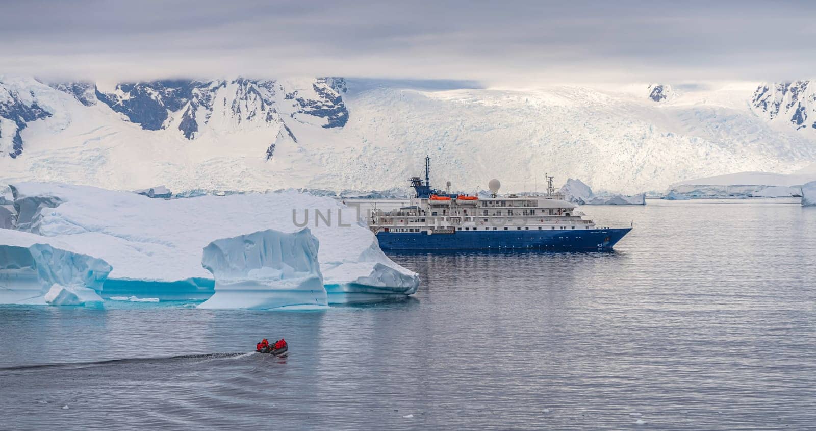 a blue, small expedition ship lies in front of an Antarctic iceberg landscape in Cierva Cove - a deep inlet on the west side of the Antarctic Peninsula, surrounded by rugged mountains and dramatic glacier fronts.