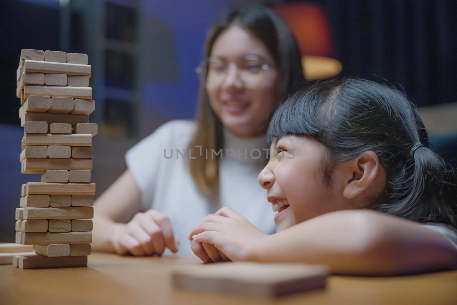 Smiling woman help teach child play build constructor of wooden blocks, Asian young mother playing game in wood block with her little daughter in home living room at night time before going to bed