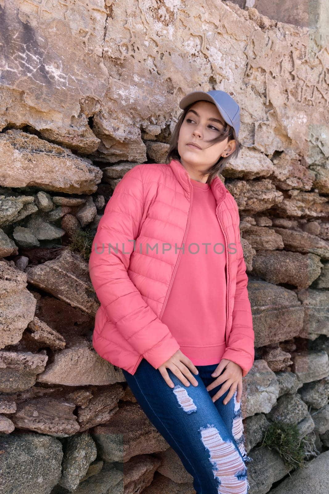Teen girl wearing pink shirt, pudded jucket and baseball cap, mockup. Serious Teenager standing outdoor near a wall and looking aside, spending her time alone. Adolescence concept