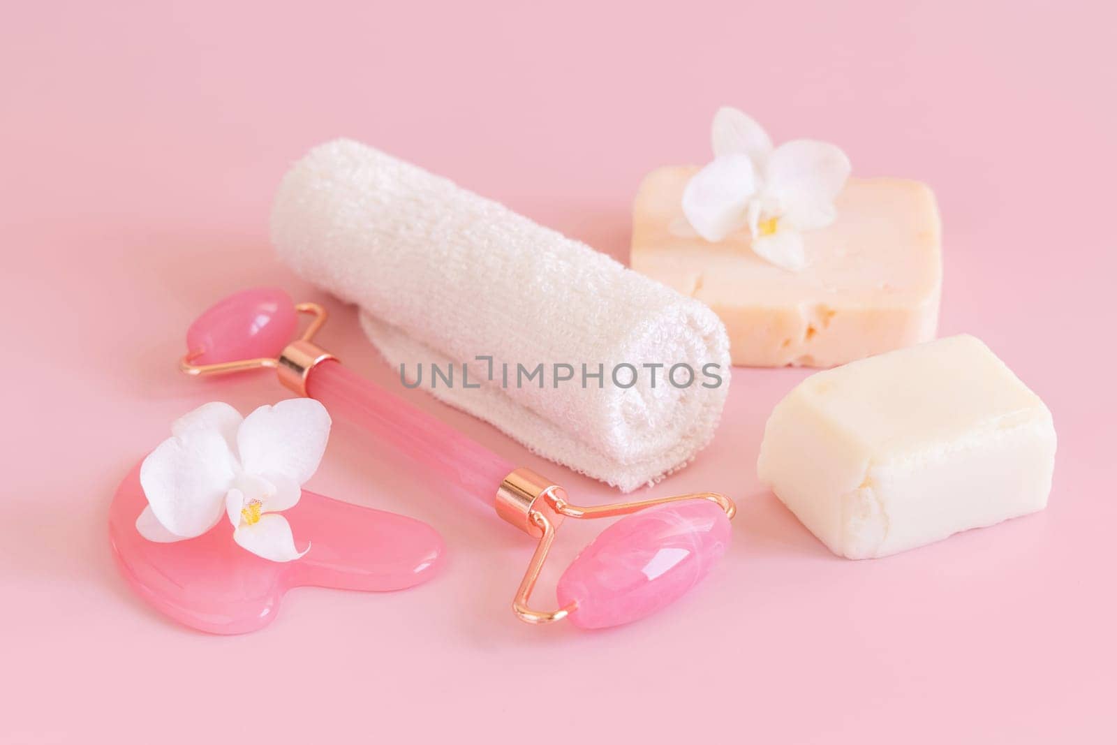 Facial roller, gua sha massager, towel and handmade soap bars near white orchid flower on pink close up. Anti-age massage and natural lifting therapy at home. Facial skin care at home. Beauty concept