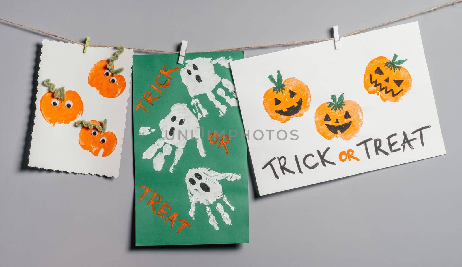 Three Halloween childrens pumpkin apple prints and ghosts handprints drawings hanging on rope on gray wall with Trick or Treat inscription. Handmade DIY decoration with children for holiday
