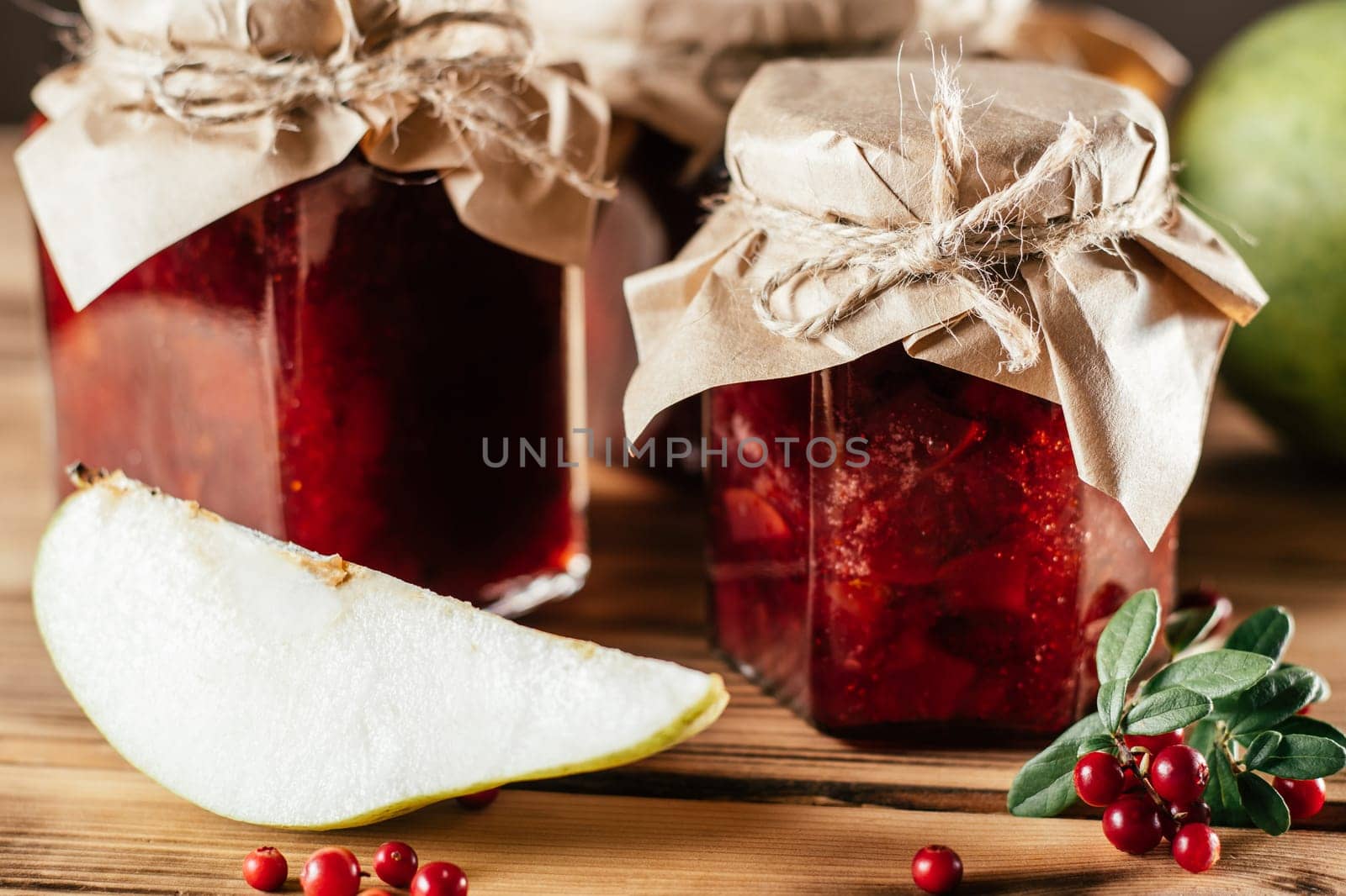 Jars of homemade lingonberry and pear jam with craft paper on lids on wooden surface next to fresh lingonberries and pears.