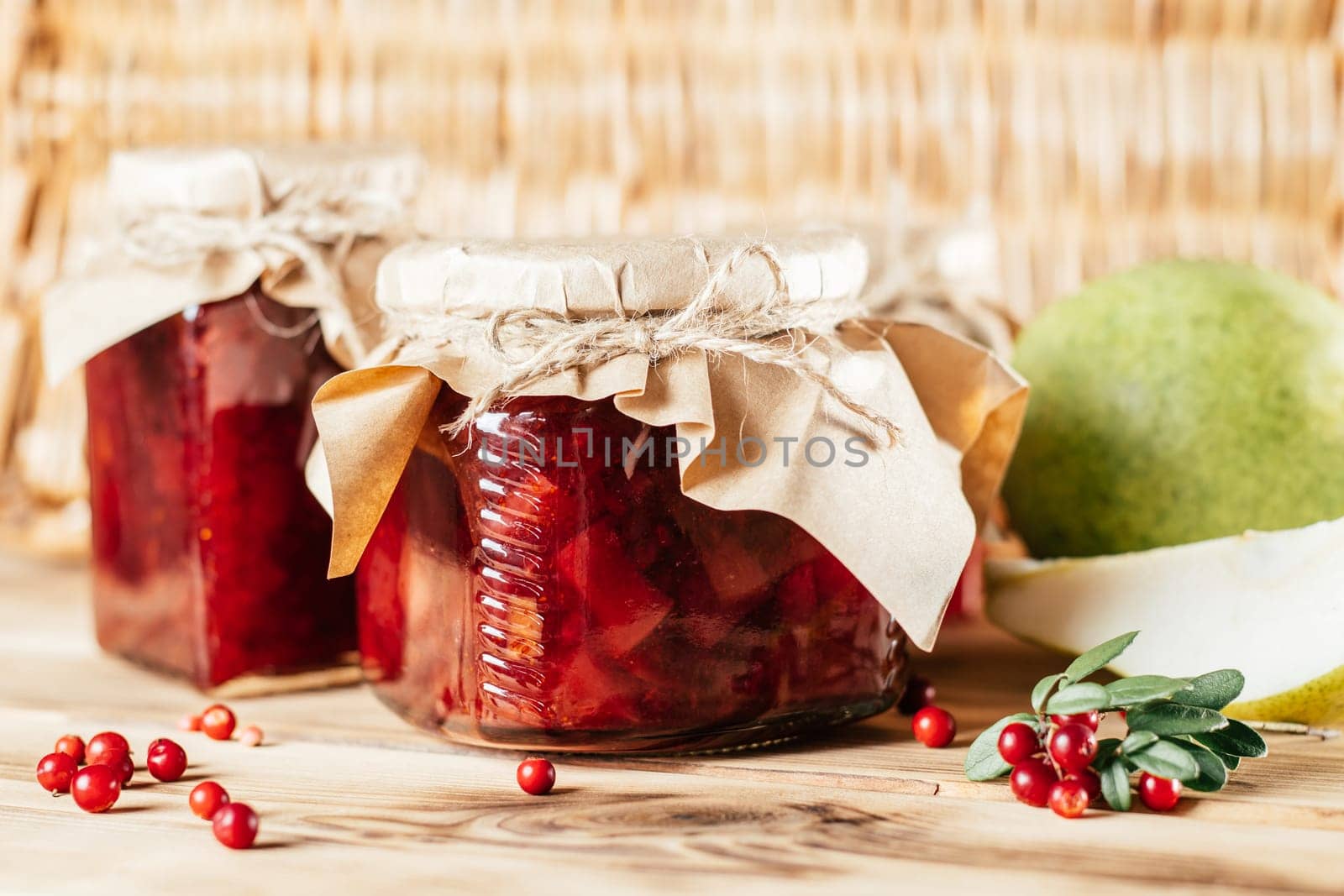 Jars of homemade lingonberry and pear jam with craft paper on lids on wooden surface next to fresh lingonberries and pears.