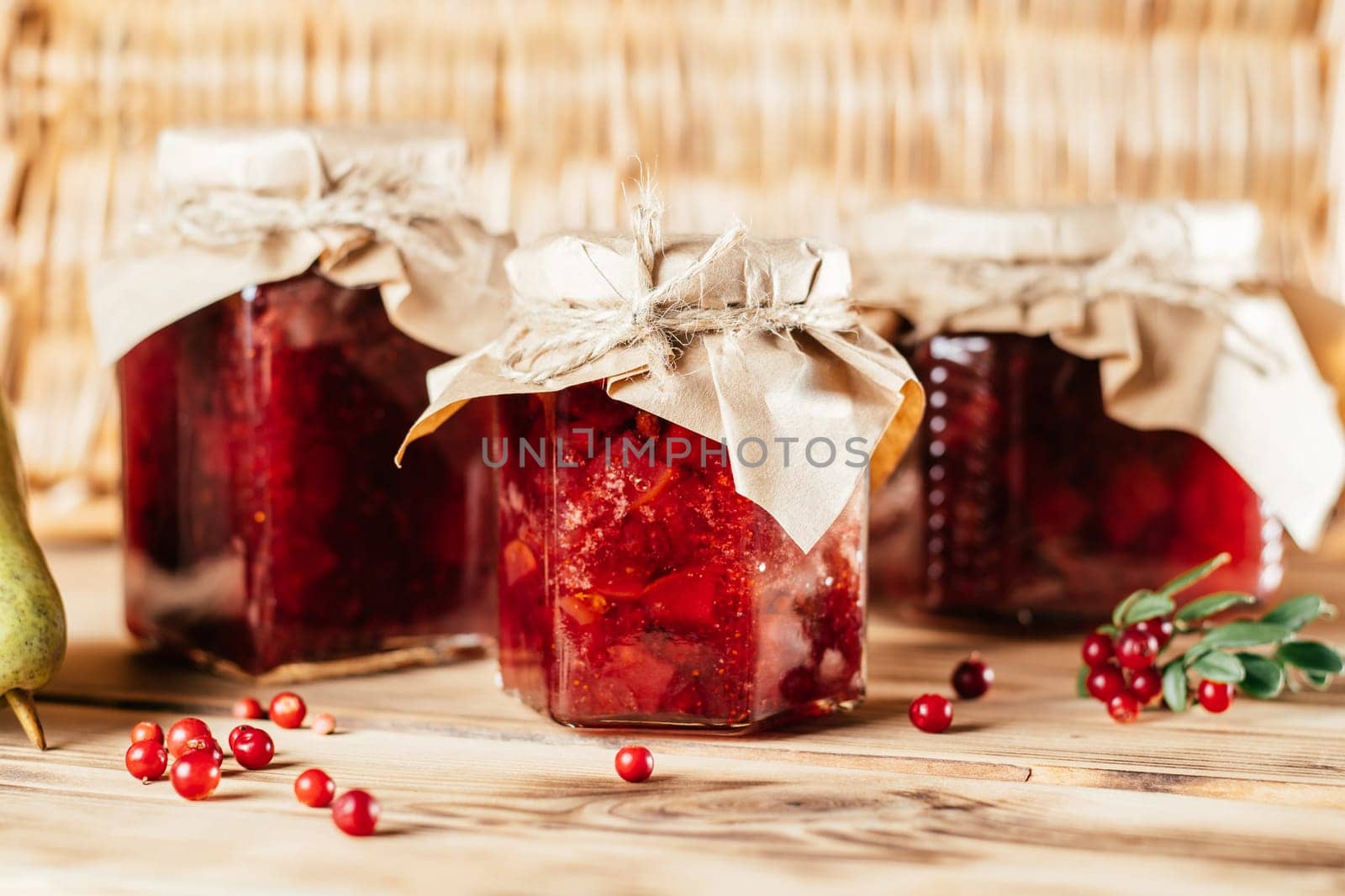 Jars of homemade lingonberry and pear jam with craft paper on lids on wooden surface next to fresh lingonberries and pears.