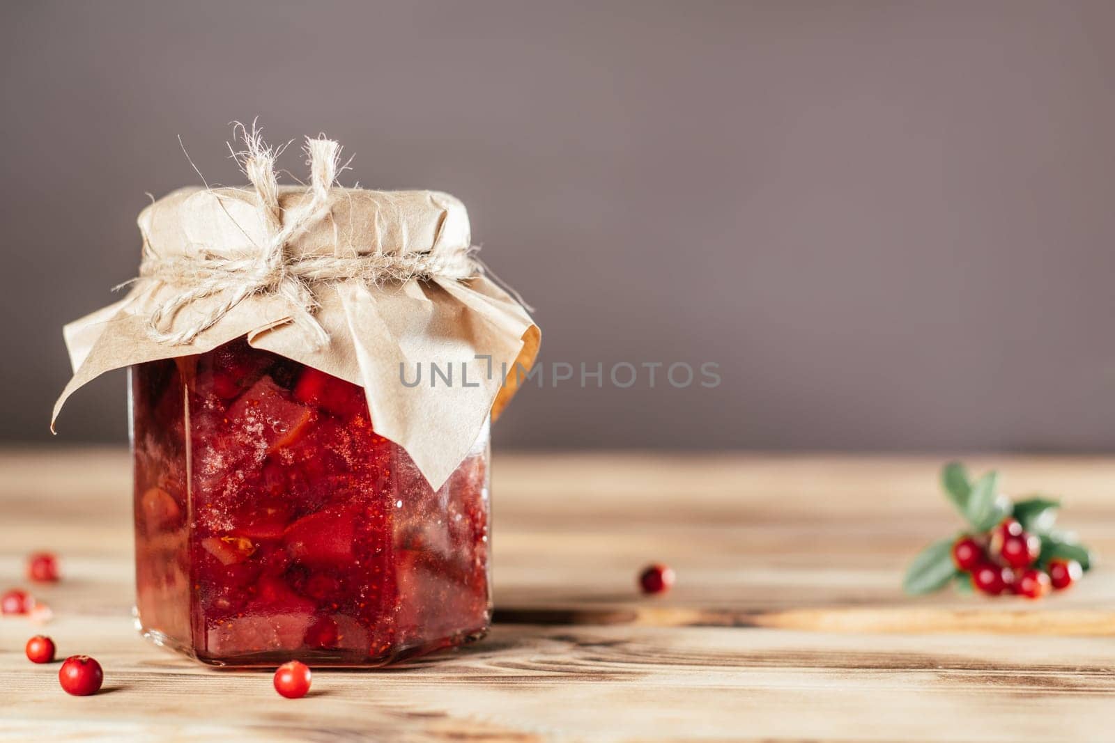 Jar of homemade lingonberry and pear jam with craft paper on lid on wooden surface next to fresh lingonberries