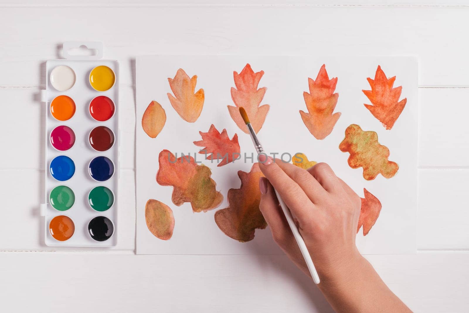 Female hand drawing autumn leaves of different shapes, sizes and colors with watercolors and brush on white wooden table. Top view. DIY handmade autumn decorations
