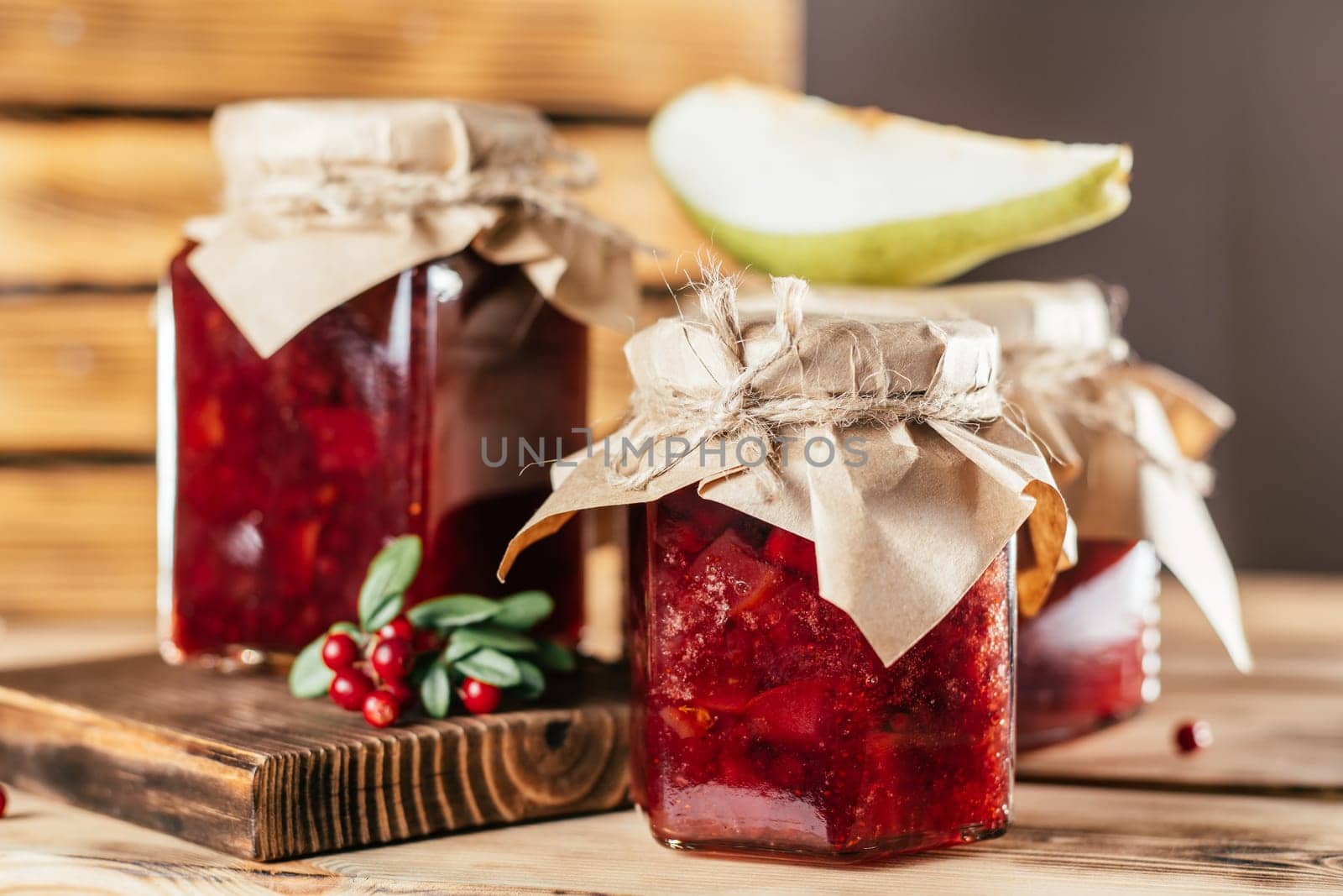 Jars of homemade lingonberry and pear jam with craft paper on lids on wooden surface next to fresh lingonberries and pears.