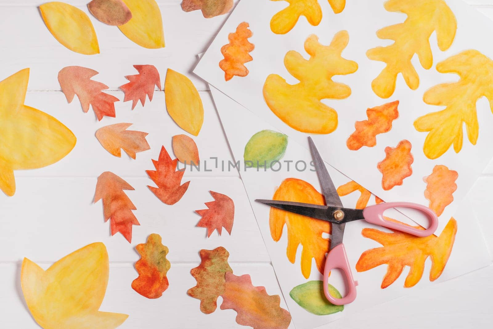 Drawn on white paper autumn leaves of different shapes, sizes and colors with scissors. Cut out leaves on white wooden table. top view. DIY handmade autumn decorations