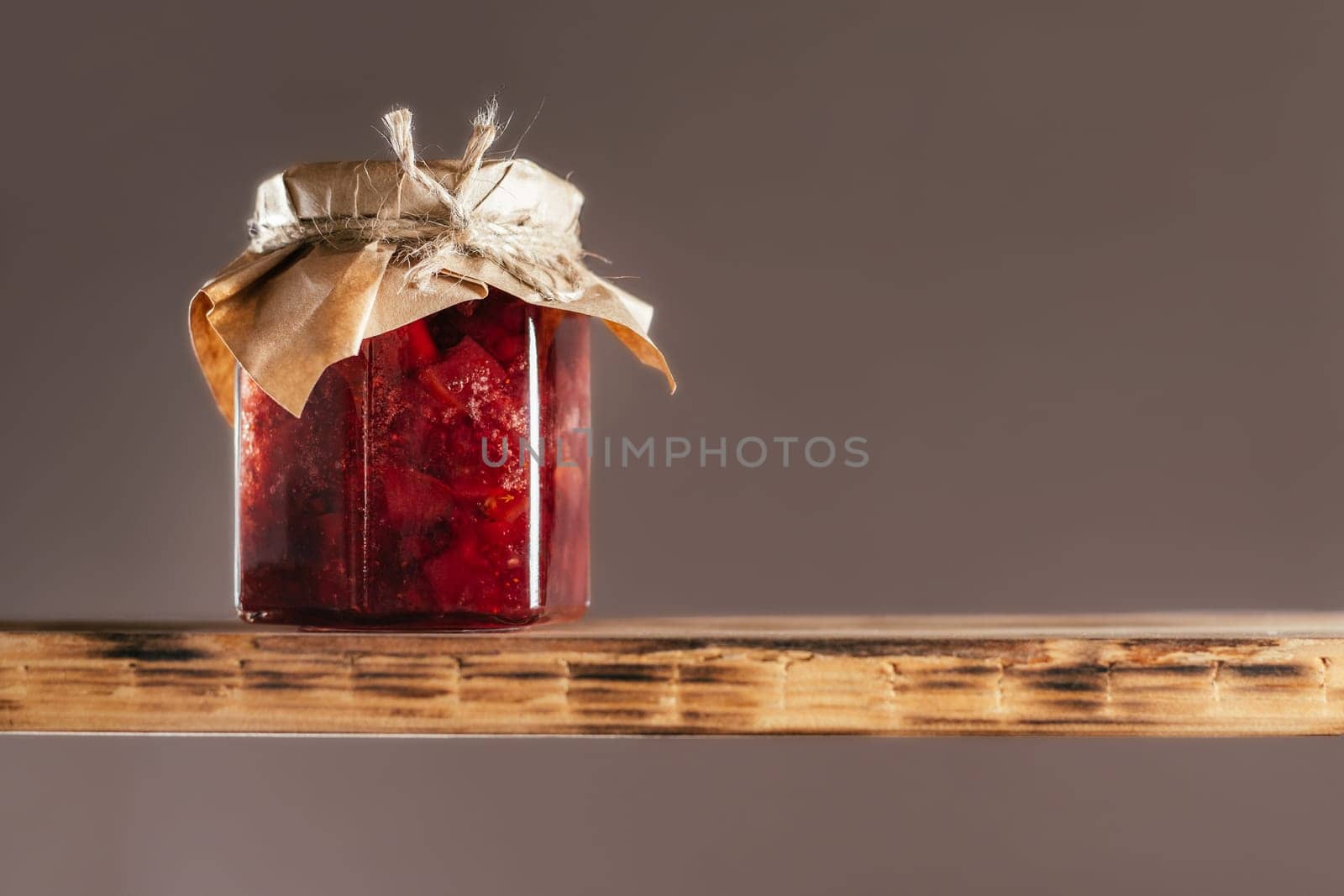 Jar of homemade lingonberry and pear jam with craft paper on lid on wooden shelf on brown background