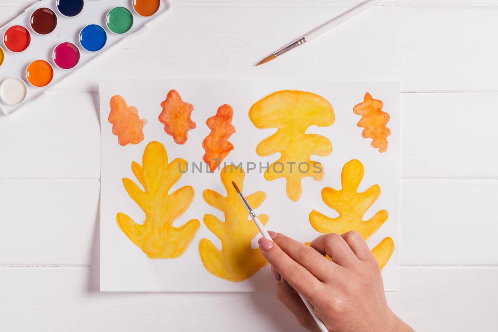 Female hand drawing autumn leaves of different shapes, sizes and colors with watercolors and brush on white wooden table. Top view. DIY handmade autumn decorations