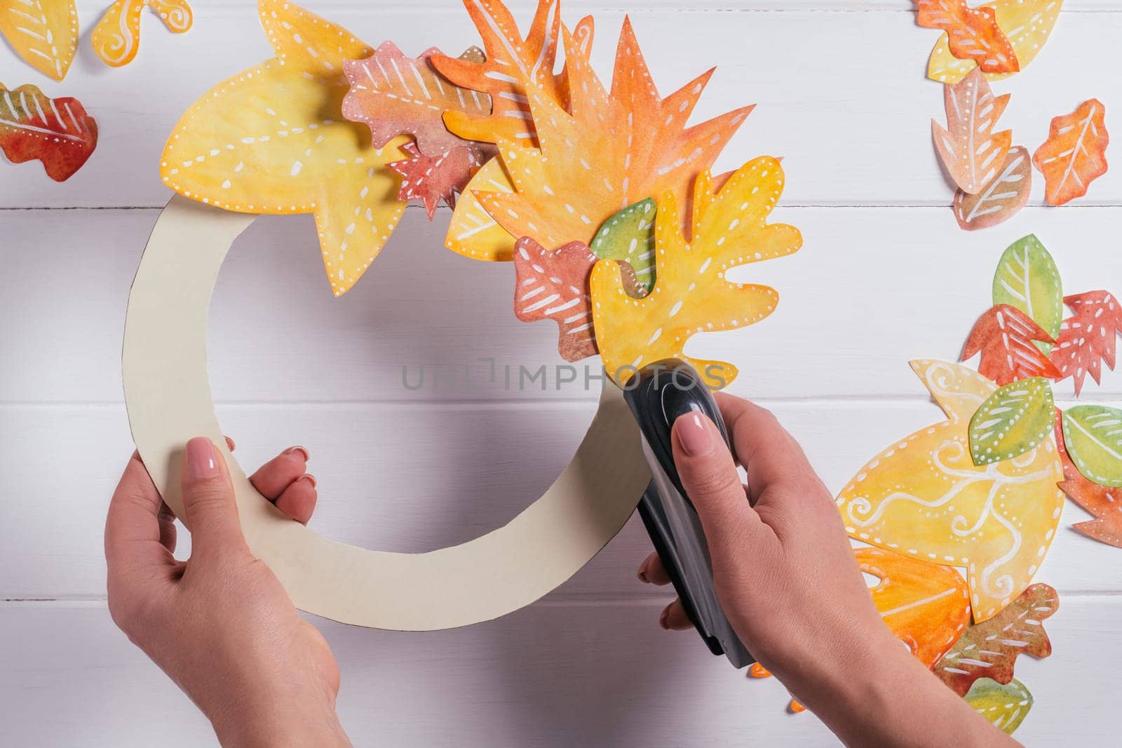 Female hands making decorative autumn paper wreath. Using stapler to fix leaves in circle on cardboard form. top view. DIY handmade autumn decorations