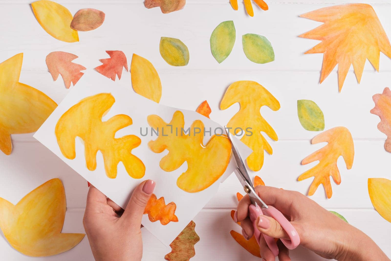 Female hands cutting out with scissors drawn on white paper autumn leaves of different shapes, sizes and colors. Cut out leaves on white wooden table. top view. DIY handmade autumn decorations