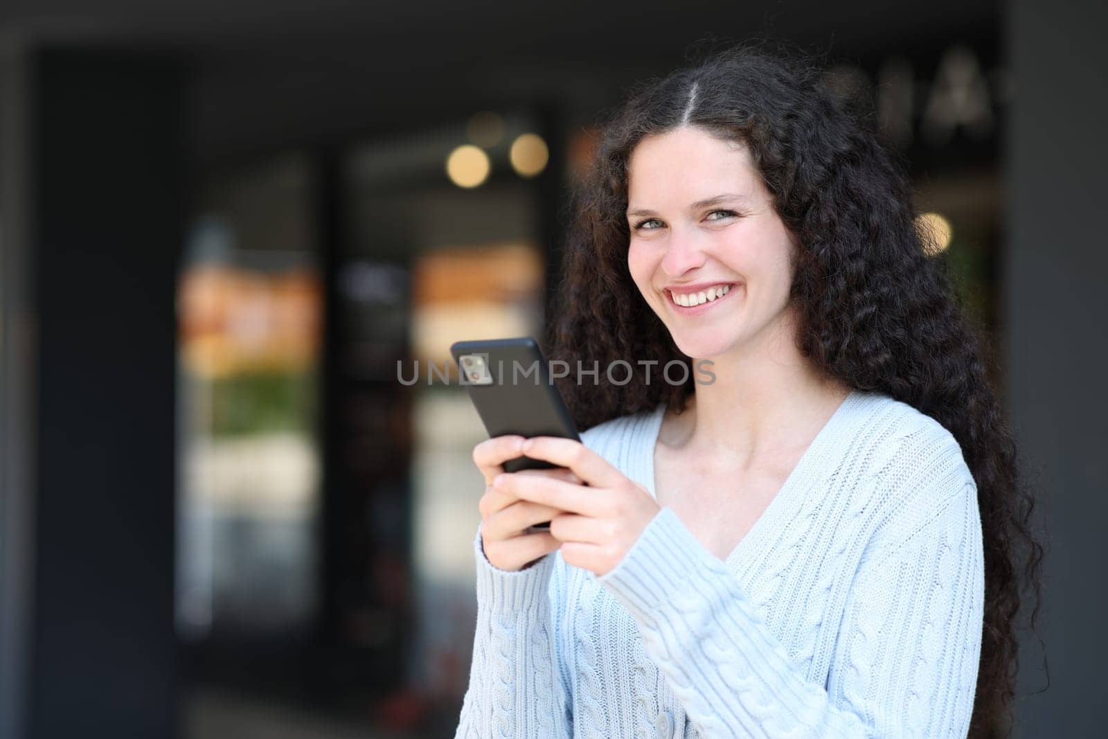 Happy woman holds smart phone and looks at camera standing in the street