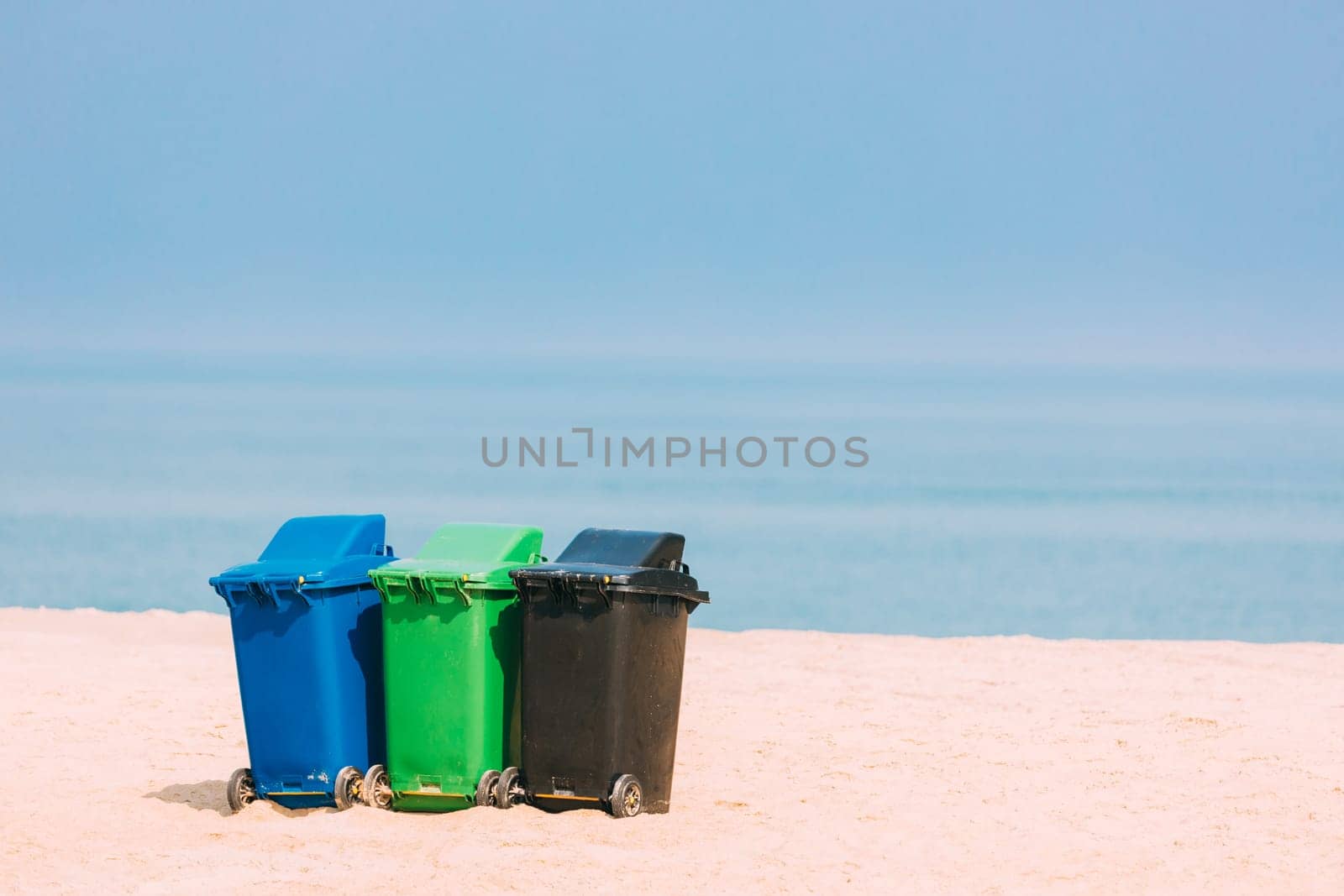 Three Plastic Waste Containers On Wheels For Separate Collection Of Garbage. Containers For Temporarily Storing Waste. Dustbins, Garbage Cans, Trash Cans On Sea Ocean Coast Beach.
