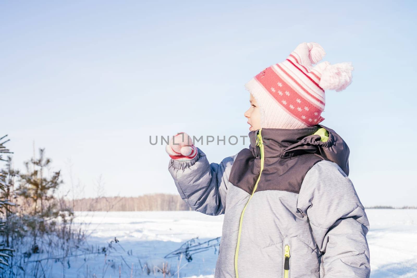 Little girl 3-4 years old in winter clothes overalls, hat and mittens, stands outdoors against clear blue sky and fir trees and looks away, pointing with hand. Winter holidays, children outdoors