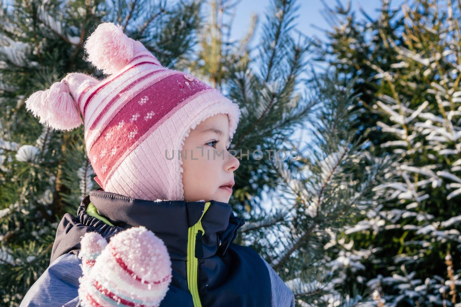 Little girl 3-4 years old in winter clothes overalls, hat and mittens, stands outdoors against snow-covered pines and fir trees and looks away thoughtfully. Winter holidays, children outdoors