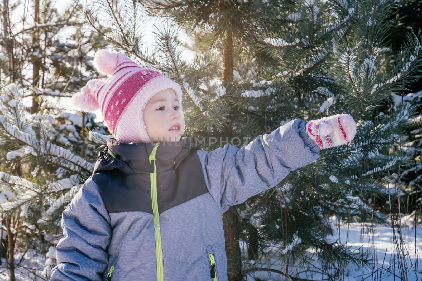 Little girl 3-4 years old in winter clothes overalls, hat and mittens, stands outdoors against snow-covered pines and fir trees and looks away, pointing with hand. Winter holidays, children outdoors