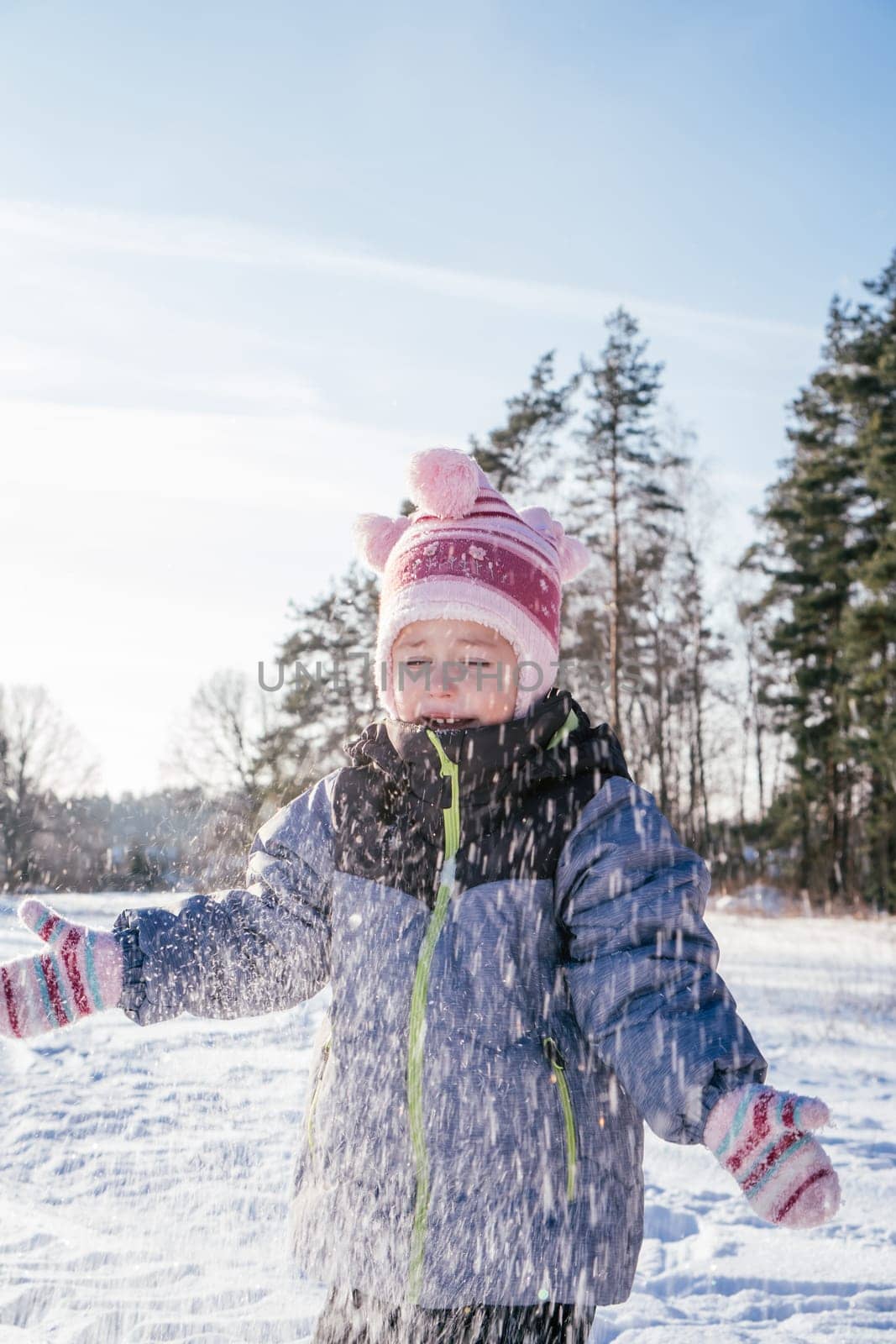 Little girl 3-4 years old in winter overalls, hat and mittens, stands outdoors against against blue sky and fir trees and throws snow over her laughing. Winter holidays, children outdoors. Vertical