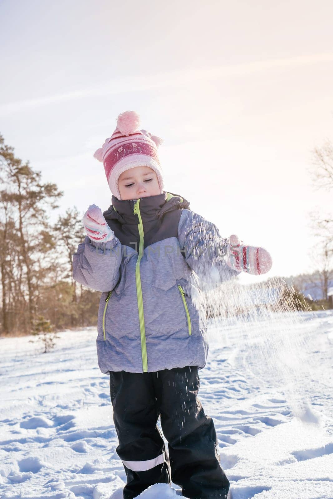 Little girl 3-4 years old in winter overalls, hat and mittens, stands outdoors against against blue sky and fir trees and throws snow over her laughing. Winter holidays, children outdoors. Vertical