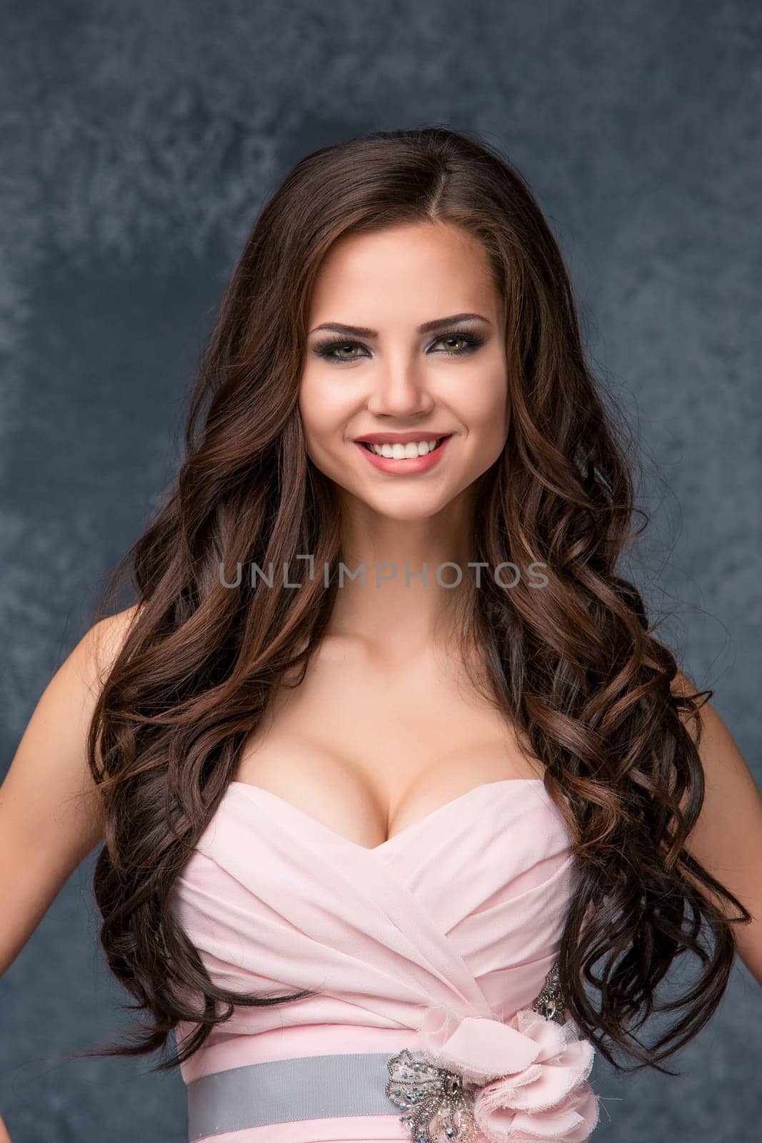 Sexy young smiling brunette woman with her hair posing in a pink dress in studio on gray background