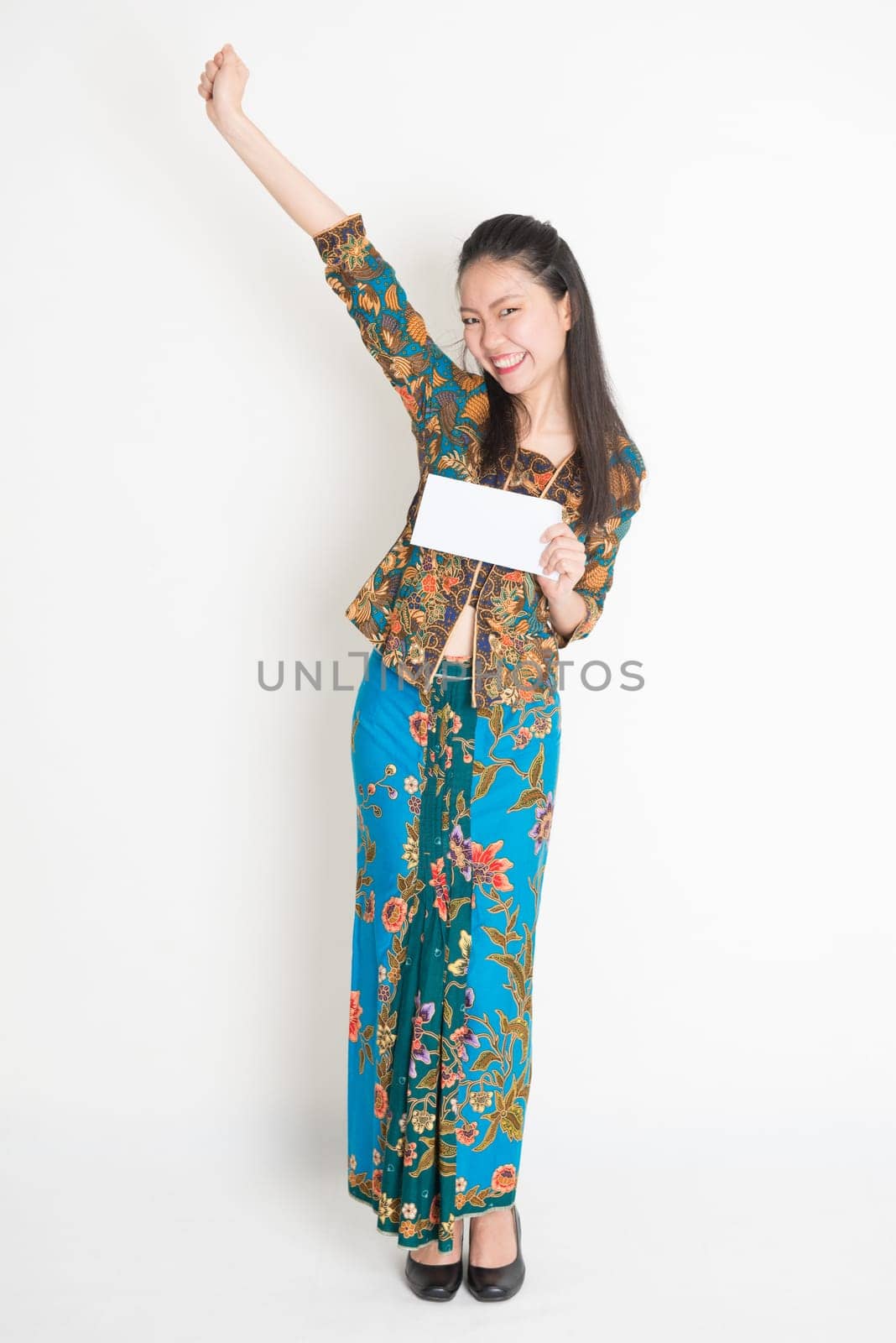 Full length portrait of Southeast Asian female in batik dress hands holding an envelope smiling, standing on plain background.