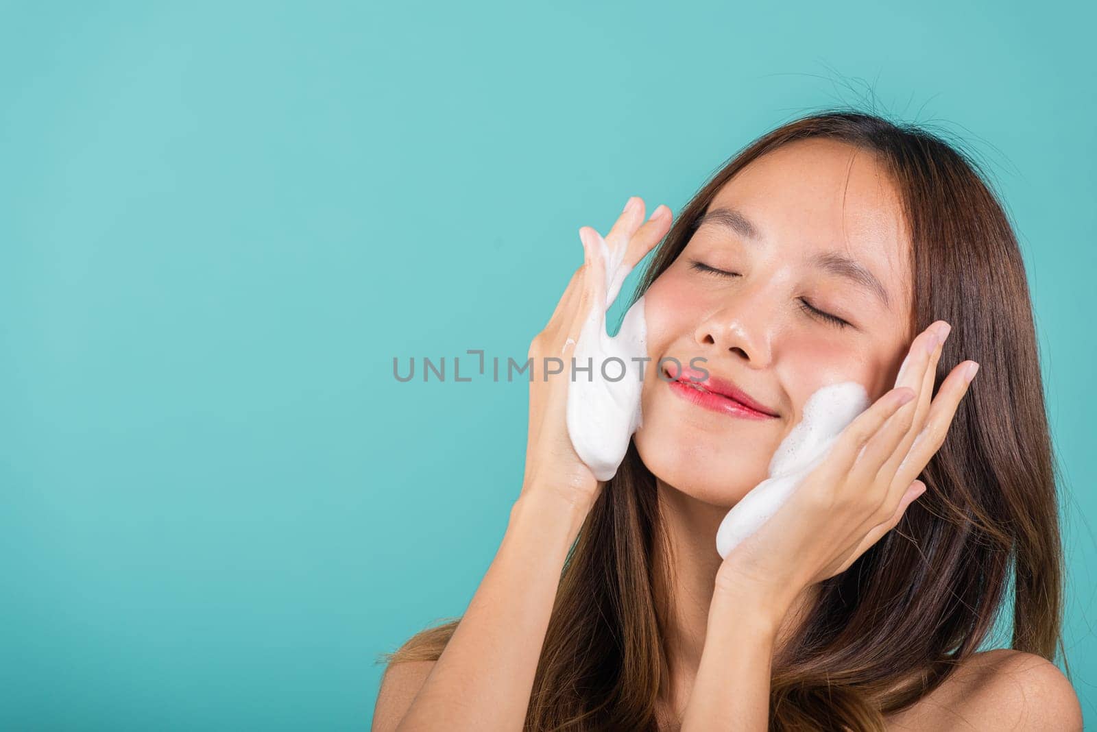 Asian woman face wash exfoliation scrub soap with skincare cleansing, Portrait beautiful female smile applies foaming cleanser healthy skin, Studio isolated on blue background, skincare daily routine