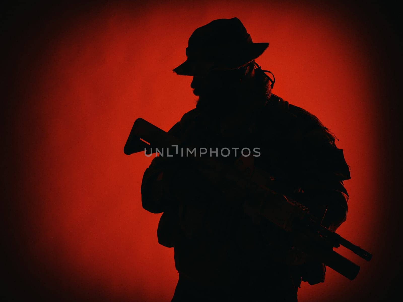 Commando shooter, army special forces rifleman in battle uniform, boonie hat, ammunition in load carrier, camouflaged with paint face, armed assault rifle, half length solhouette portrait on red background