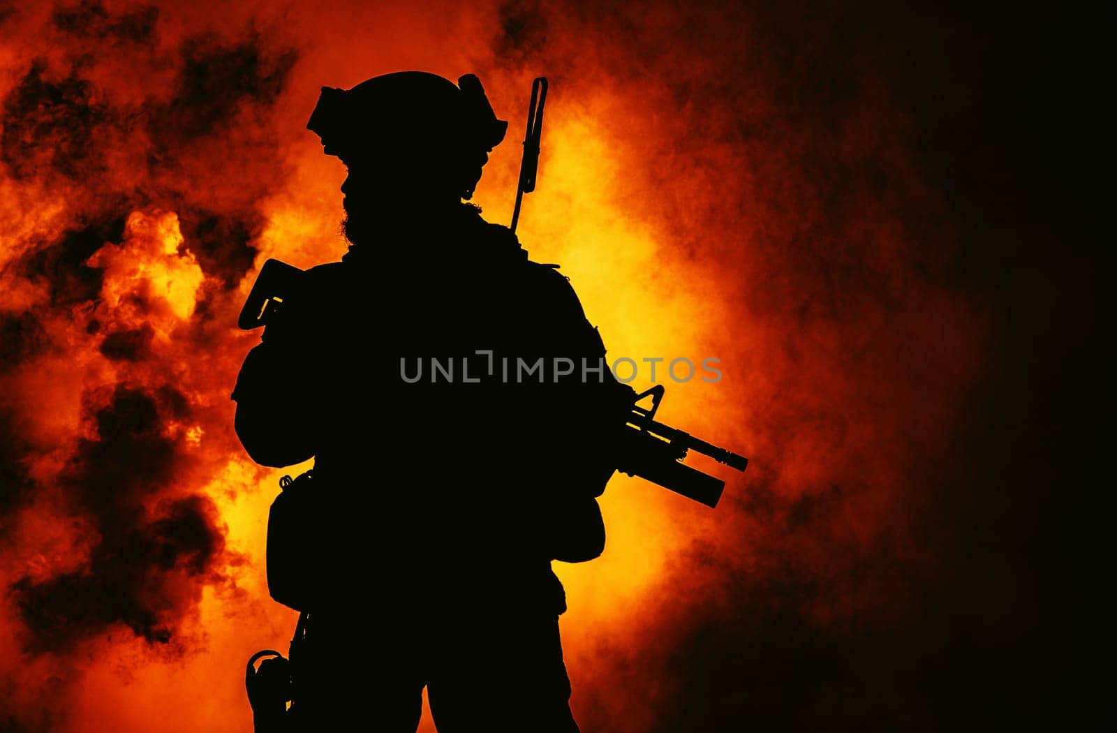 Silhouette of modern infantry soldier, elite army fighter in tactical ammunition and helmet, standing with assault service rifle in hands in the fire and smoke