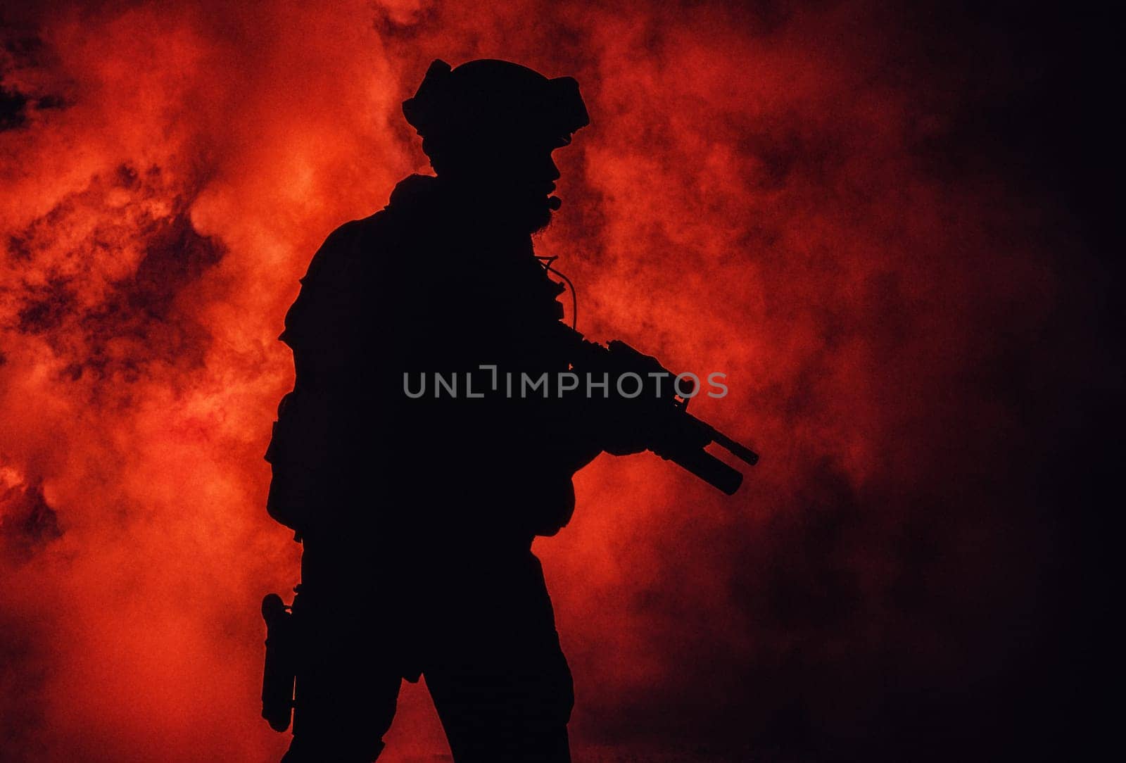 Silhouette of modern infantry soldier, elite army fighter in tactical ammunition and helmet, standing with assault service rifle in hands in the fire and smoke