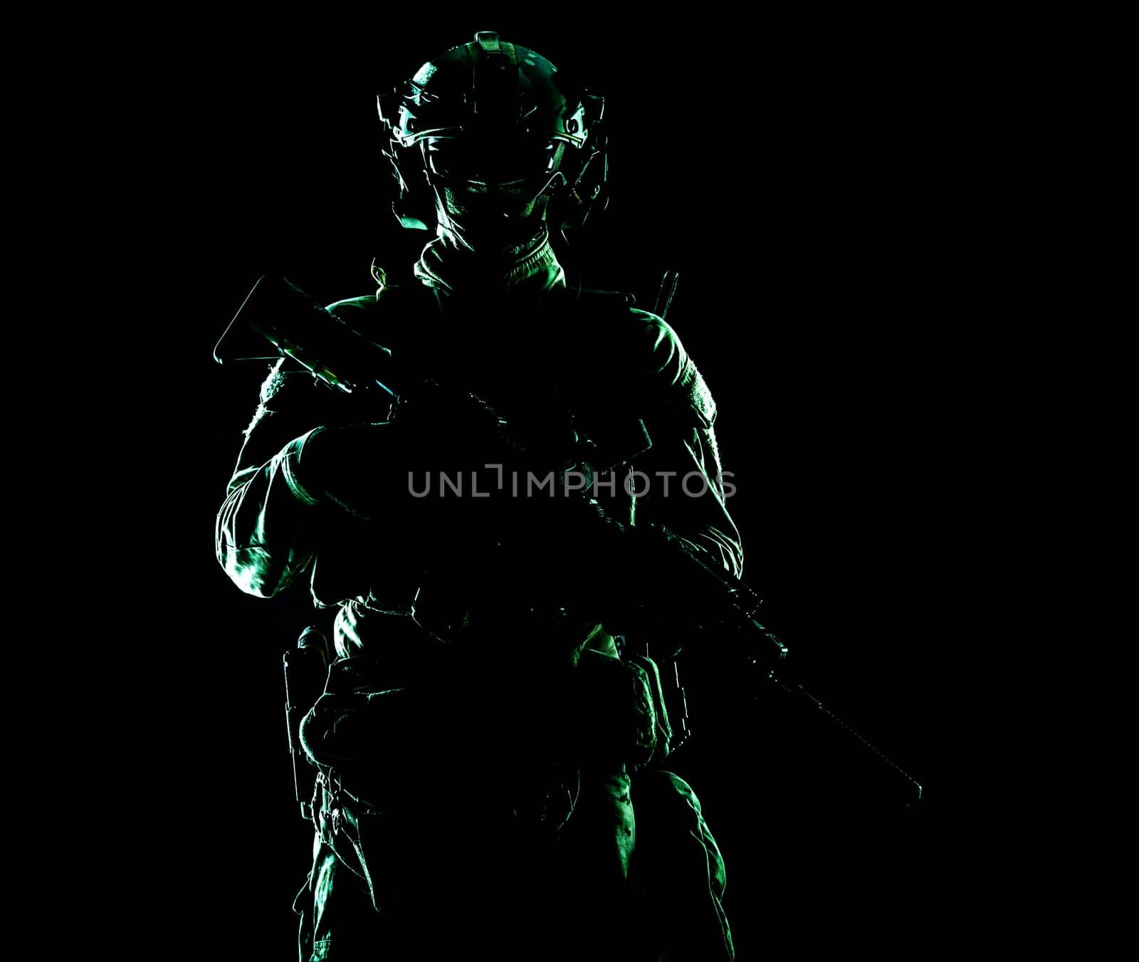 Army special forces elite soldier with hidden behind mask and glasses face, battle helmet, tactical radio headset, standing with assault rifle equipped silencer in darkness, contour shot