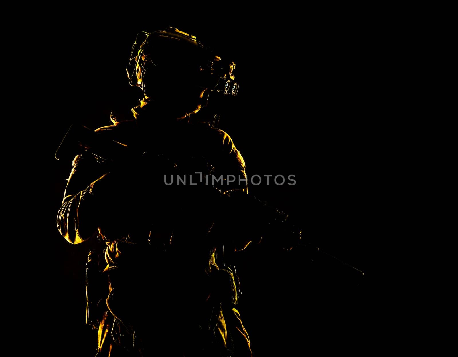Army special forces elite soldier with hidden behind mask and glasses face, battle helmet, tactical radio headset, standing with assault rifle equipped silencer in darkness, contour shot