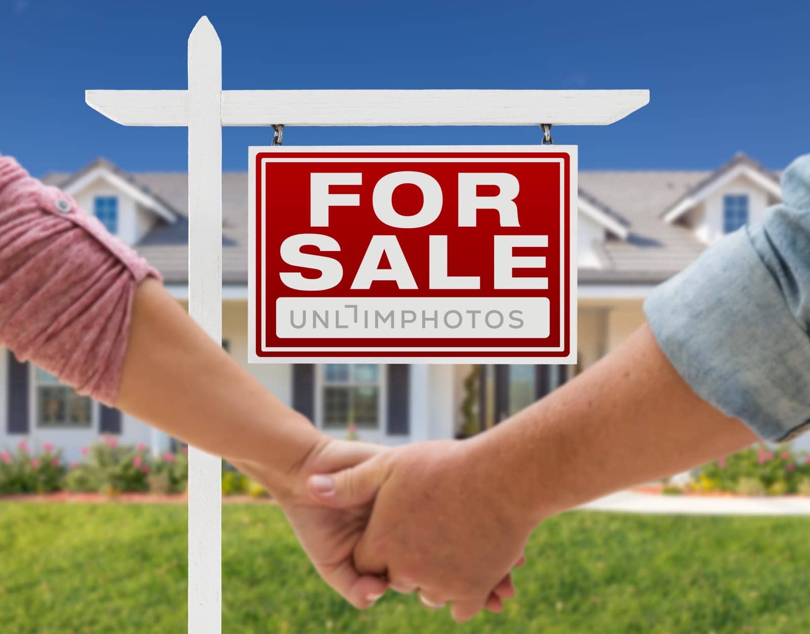 Couple Holding Hands In Front of Beautiful New House and For Sale Real Estate Sign.