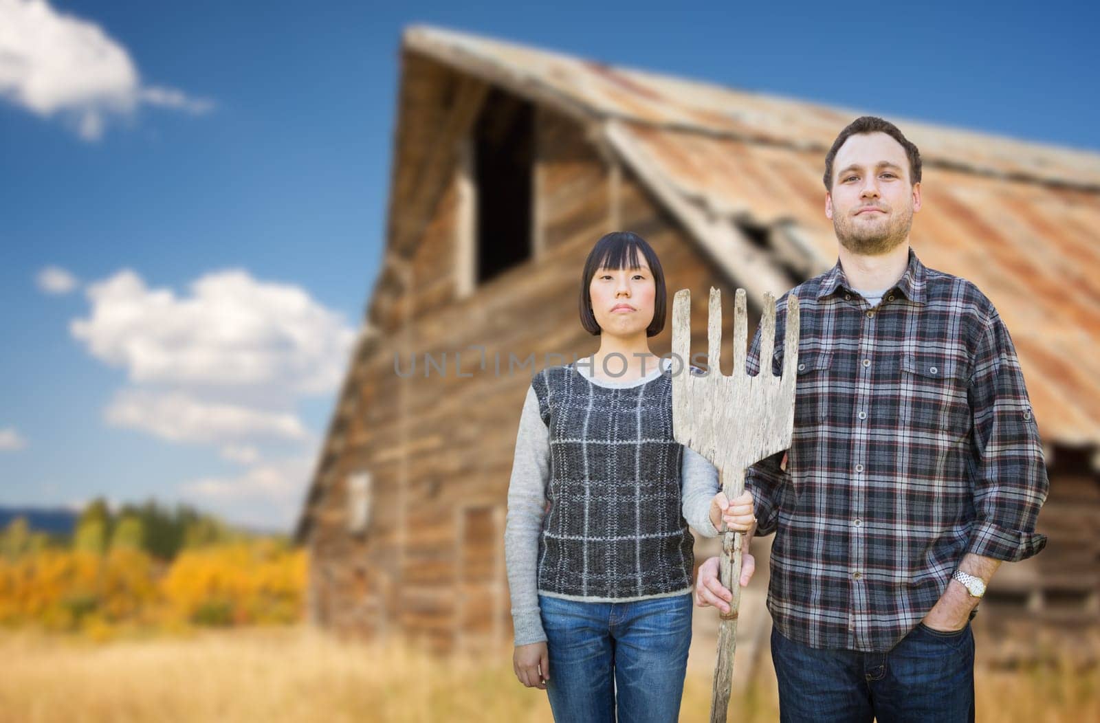Biracial Chinese and Caucasian Young Couple Holding Wooden Pitch Fork In Front of Rustic Barn In The Country.