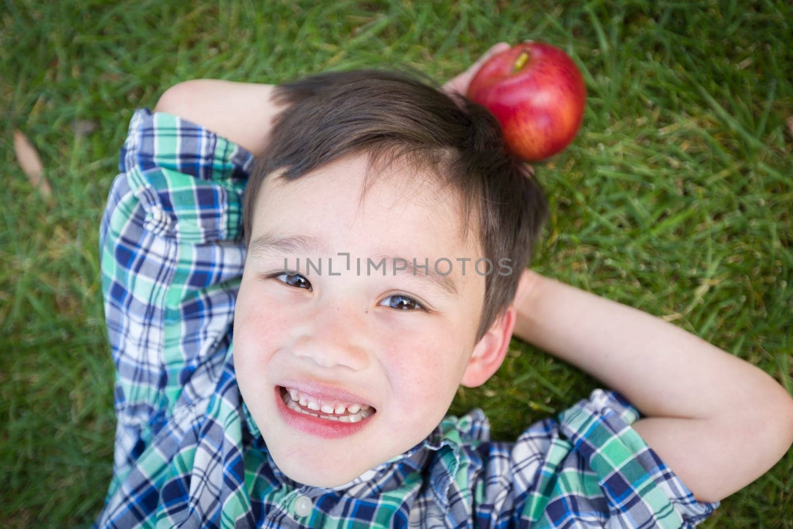 Mixed Race Chinese and Caucasian Young Boy With Apple Relaxing On His Back Outside On The Grass.
