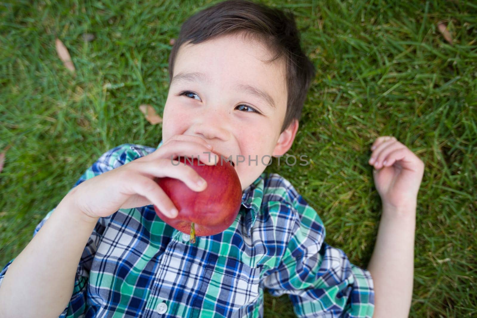 Mixed Race Chinese and Caucasian Young Boy With Apple Relaxing On His Back Outside On The Grass.