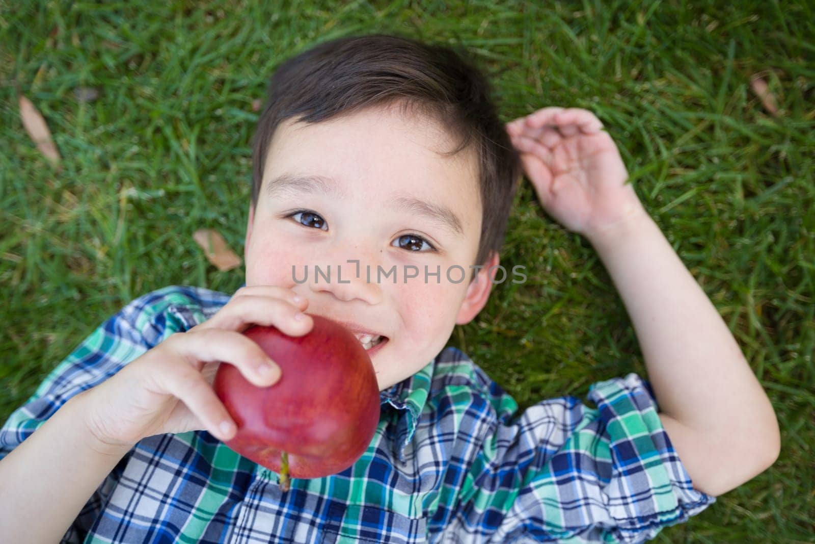 Mixed Race Chinese and Caucasian Young Boy With Apple Relaxing On His Back Outside On The Grass.