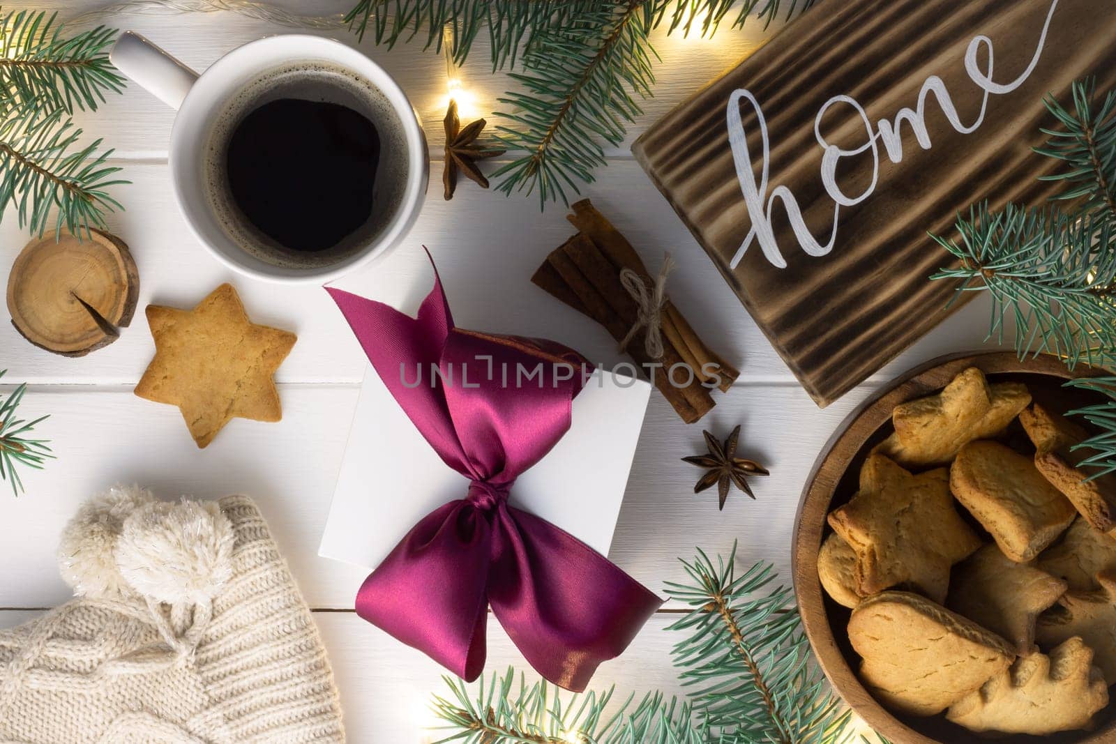 Home lettering burnt wood sign, gingerbread cookie, Christmas tree branches, cup of coffee, gift, cinnamon sticks on white surface. Flat lay