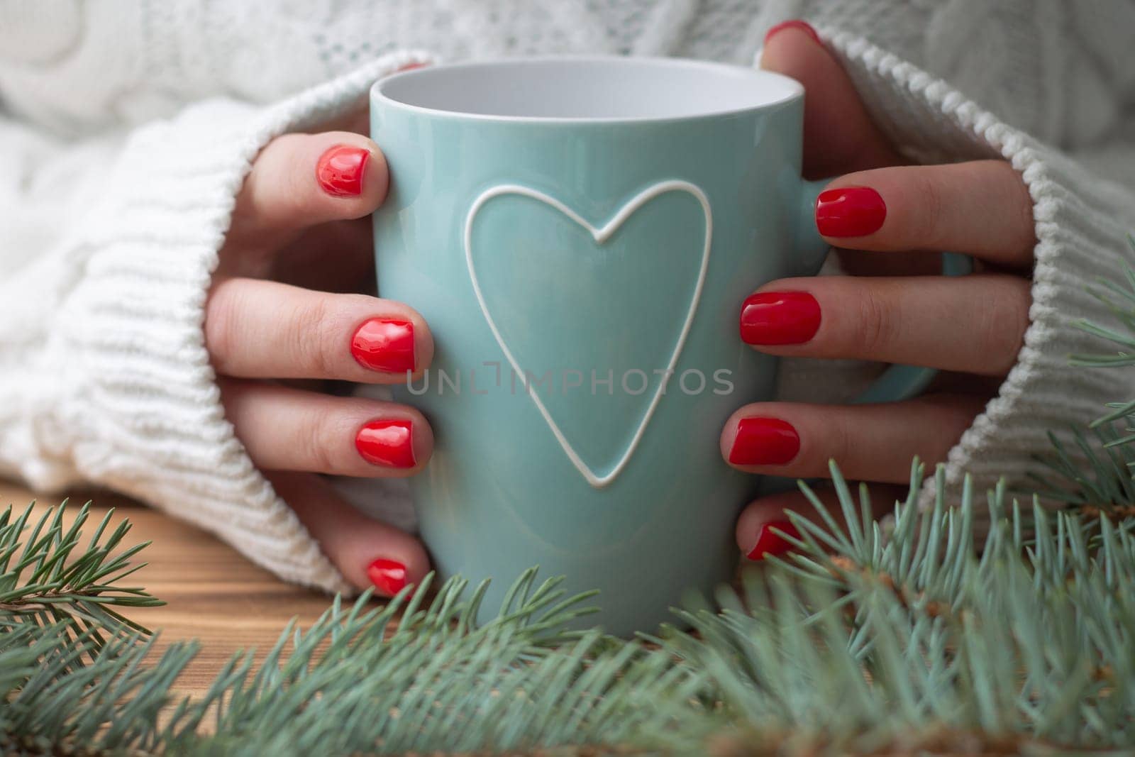 A woman's hands in a white knitted sweater with red nail polish hold a mint mug with a heart, near the branches of a Christmas tree.
