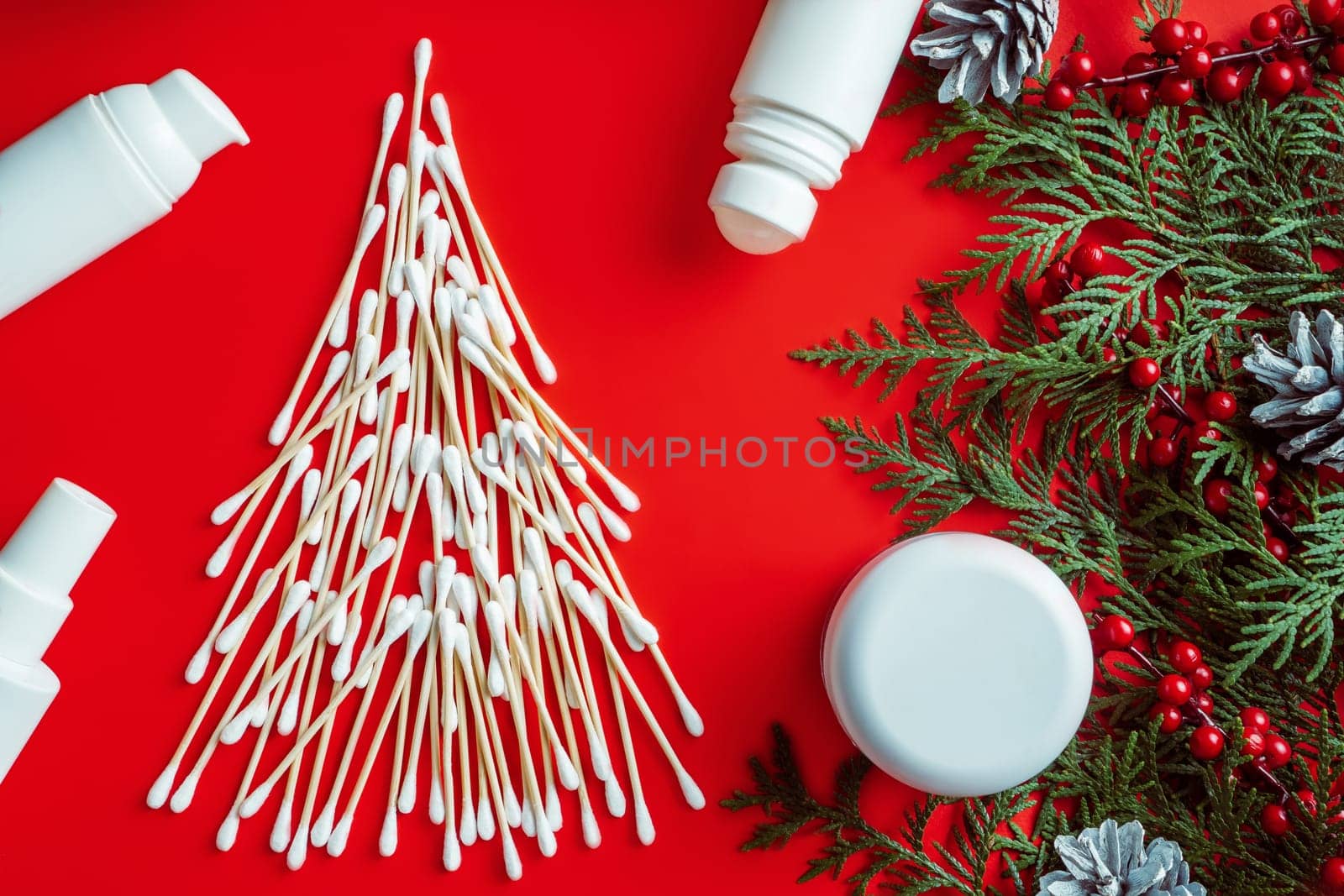 Wooden cotton swabs laid out in shape of Christmas tree with cosmetics jars, roll-on deodorant and spruce branches, cones, red berries on red background. Flat lay