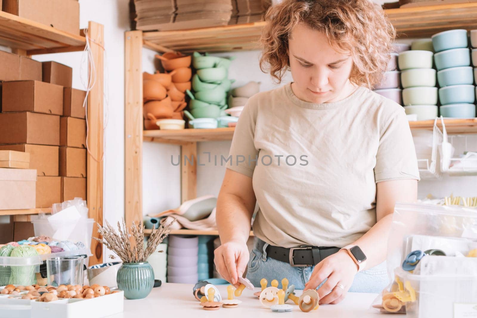 Blond confident curly woman making and design craft silicone pacifiers for newborn babies on white desk. Small business, store. Choosing soft material for kids. Natural ecological accessories