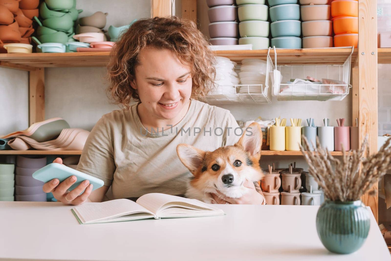 Smiling concentrated businesswoman holding smartphone, with cute calm dog corgi in workshop. Set of silicone baby dishes near wall on shelves. Home office, taking orders in comfortable atmosphere