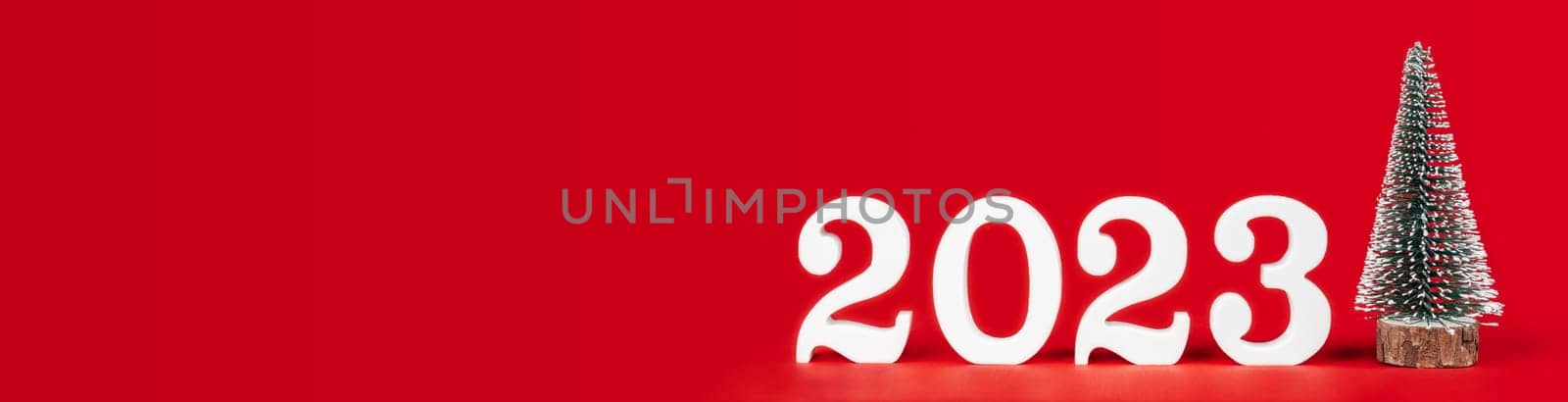 Happy New Year 2023. Bright red long rectangular banner with white wooden numbers 2023 stand with toy Christmas tree. Merry Christmas. Copy space. Place for text