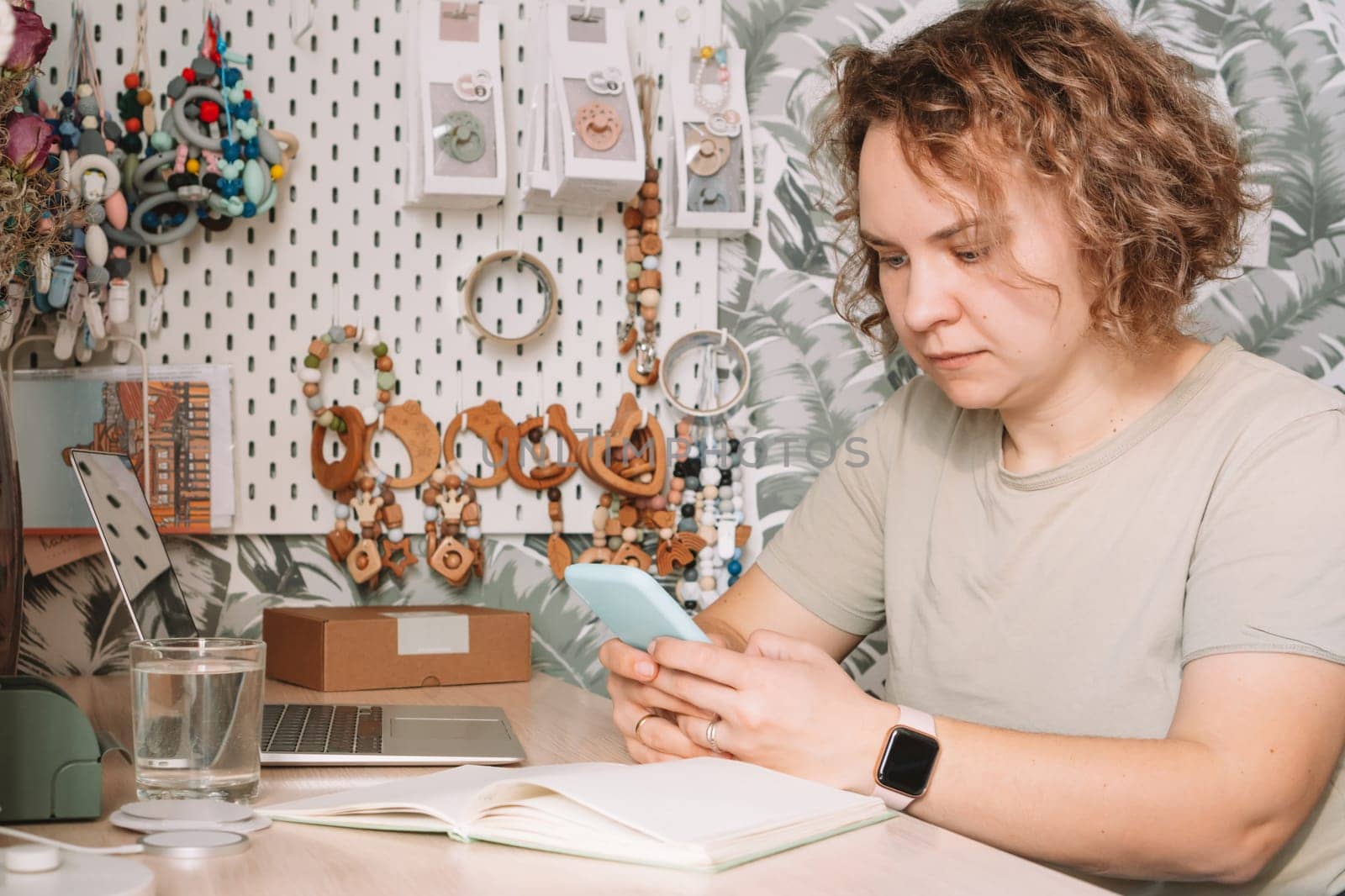 Smiling concentrated business woman talking smartphone, working on laptop in workshop. Baby healthcare accessories, silicone eco rattles, teethers, pacifiers, hanging on white wall. Home office
